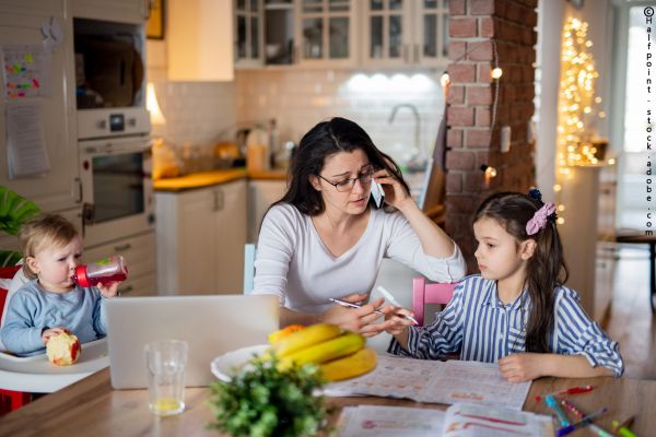 Mutter sitzt mit ihren Kindern am Küchentisch und arbeitet, telefoniert und übernimmt die Kinderbetreuung gleichzeitig